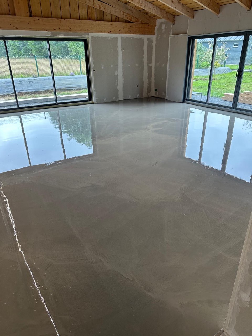 Dalle béton lissée fraîchement coulée dans une pièce à grandes baies vitrées et charpente bois apparente — chantier à Genneville
