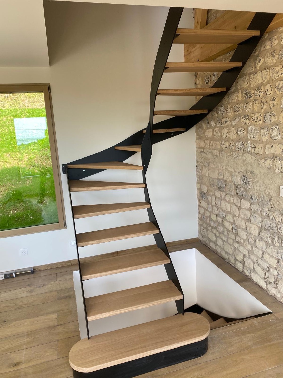 Escalier sur mesure à limon métal noir et marches en chêne contre un mur en pierre apparente