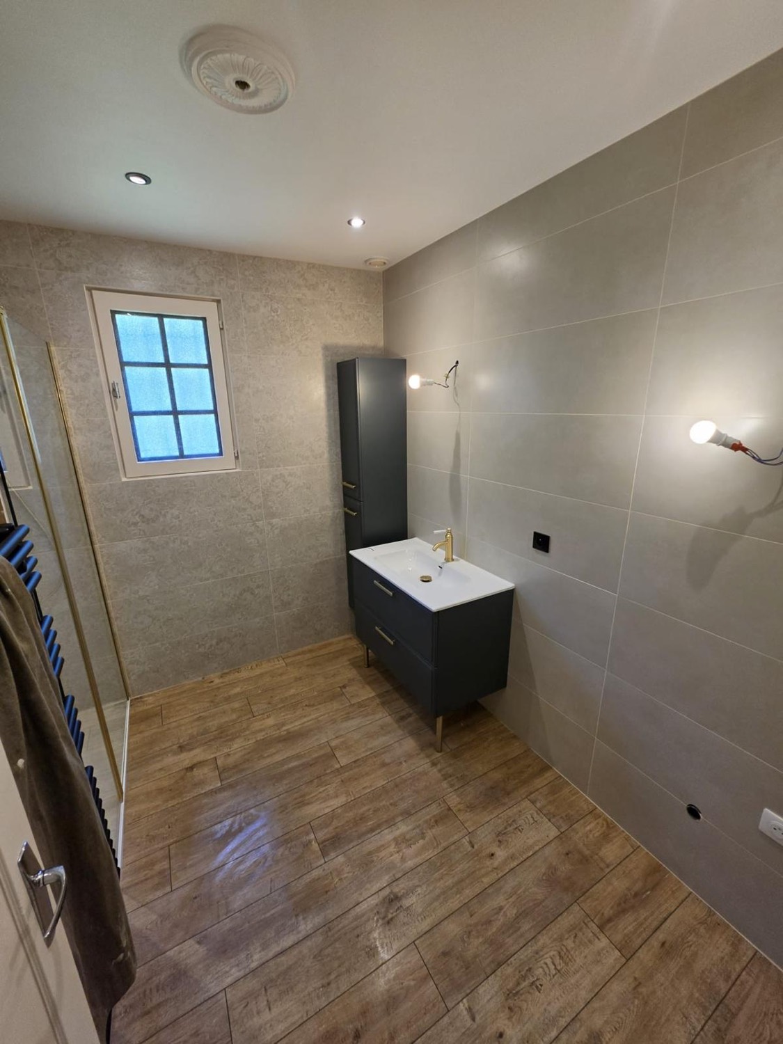 Salle de bain avec meuble vasque bleu pétrole, robinetterie laiton, colonne de rangement et sol carrelage effet bois